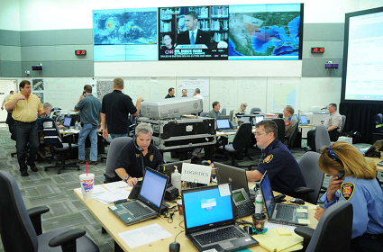 Emergency Operations Center bustles with activity, with some personnel seated at desks with laptops and speaking on telephones, and others standing near the desks.  Three large projection screens display different news channels on a wall.