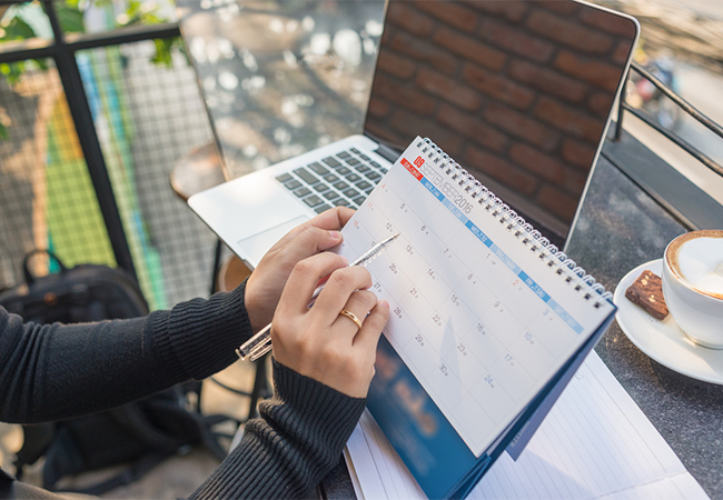 A woman's hand pointing at a calendar with a pen, with a laptop in the background.