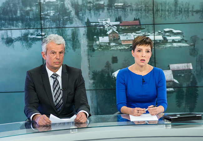 Two news reporters with an image of a flooded town on a screen behind them.