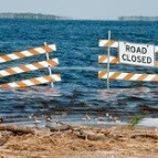 Photograph of a lake that has flooded and gone beyond two closed road barriers, one with a Road Closed sign attached