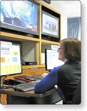 Woman working at computer in front of television screens showing weather forecasts in an Emergency Operations Center