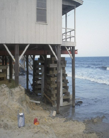 The image is a photo of the lower corner of a house adjacent to the ocean. The house is elevated on pilings and on stacked pallets. the ocean encroaches the pilings. It appears that the beach on which the house sits has been eroded.