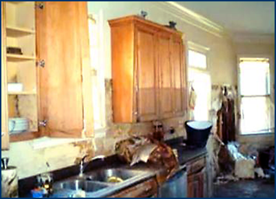 Photo of one side of a kitchen showing widespread major damage to both base and wall cabinets, as well as counters