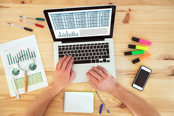 Person on a computer working on a spreadsheet with documents and pens nearby.