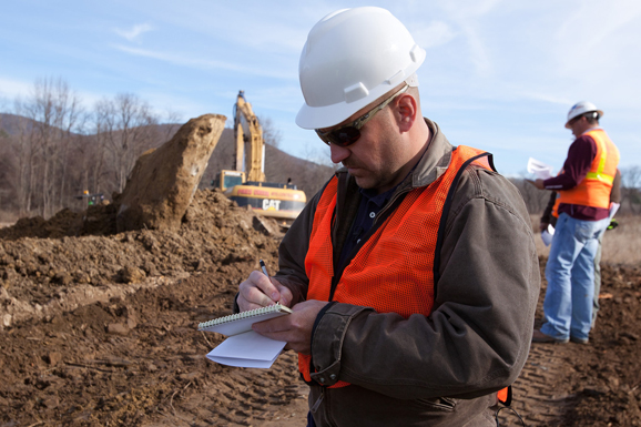 Image of someone in a safety vest and hard hat taking notes at a work site