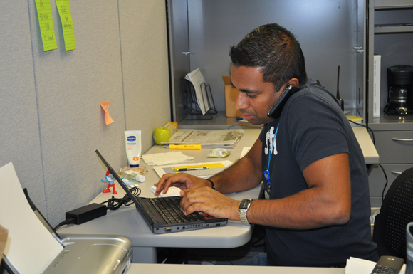 A person working on a laptop and talking on a mobile phone in a cubicle.