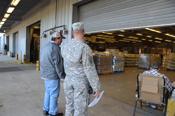 View of pallets in a warehouse through a partially-open loading door. A National Guard officer is speaking to a truck driver in the foreground.