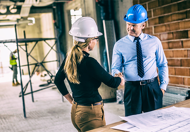 An architect shaking hands with a businessperson over plans at a construction site.