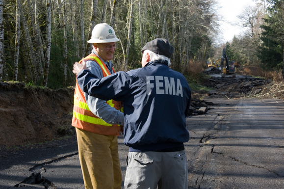 In the middle of a damaged road, someone in a FEMA jacket shakes hands with someone wearing a safety vest and hard hat.