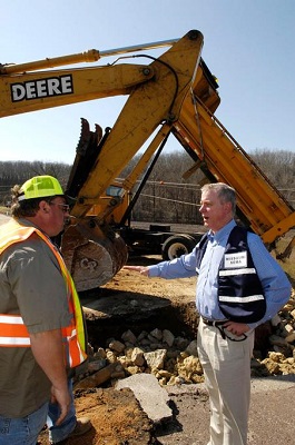 An official speaking with a road worker in front of a backhoe at a construction site.