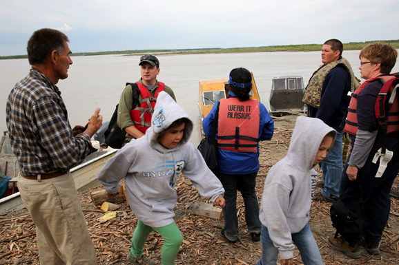 A FEMA inspector meeting with a disaster survivor and his grandchildren, who play on the bank of the Yukon river.