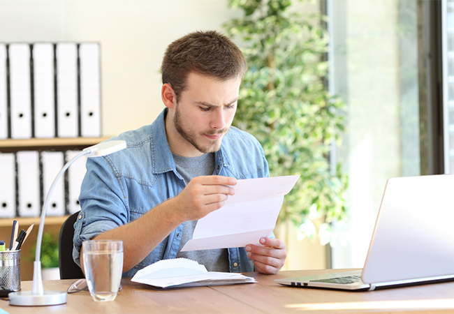 A man sitting at a desk in front of a laptop, opening a letter.