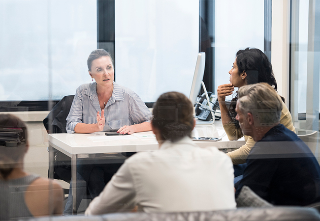 A business woman sitting at a desk, speaking to four other people at a meeting.