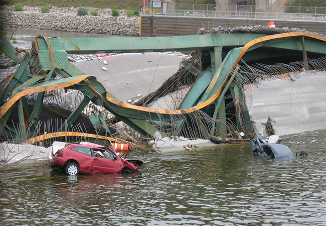 Cars and roadway litter the river under a collapsed bridge.