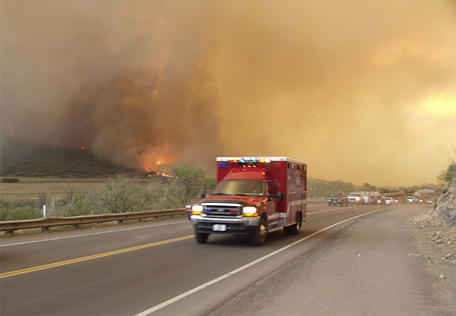 Evacuees leaving town during wildfire outbreak with an emergency vehicle at the front.