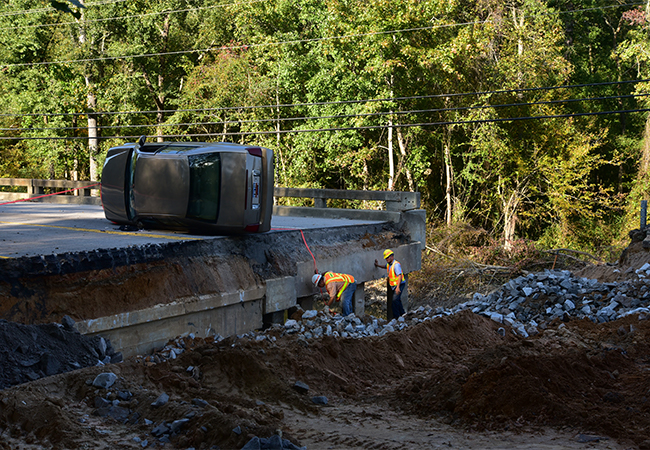 Vehicle on its side on a bridge damaged by a flood with transportation workers digging through debris.