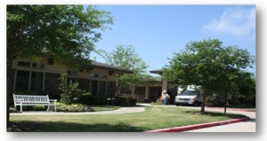 A photo of an assisted living facility on a tree lined street.  A facility bus is parked under a portico, and there is a bench in front of a walkway.