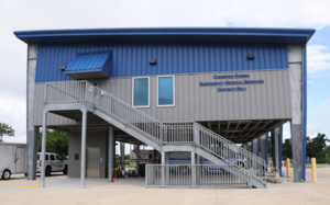 A photo of a raised metal building with the words Cameron Parish Emergency Medical Services District Office on the front of the building. A staircase leads from the ground level up to the front door.  Several vehicles are parked underneath the building.