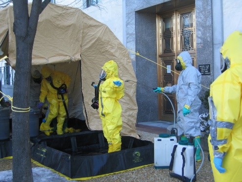 Responders in Level A personal protective equipment suits conducting decontamination activiites including spraying down a responder at center.